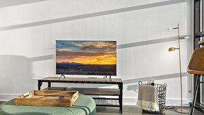 A modern living room with a flat-screen TV on a wooden stand, a green ottoman, a woven basket, and a tall floor lamp.