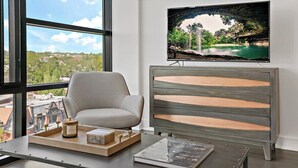 Une chambre moderne avec une grande fenêtre, un fauteuil gris, un meuble en bois, un téléviseur à écran plat et un plateau avec des bougies et des livres.
