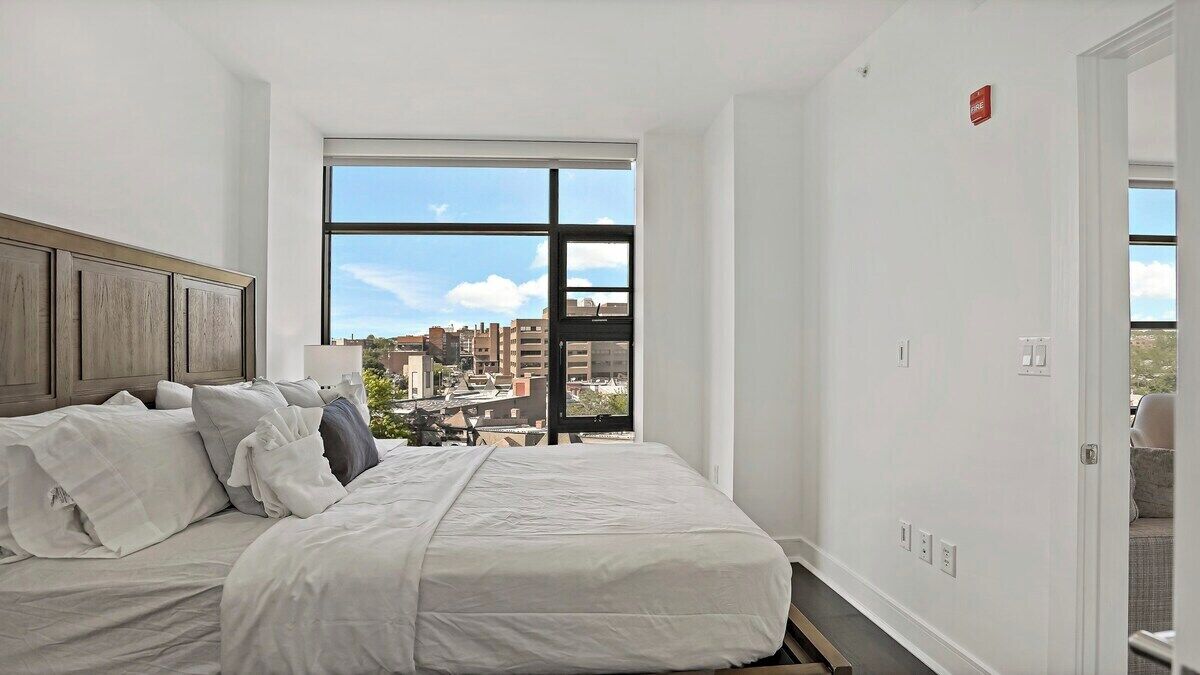 A bedroom with a large window, a bed with white bedding, and a view of buildings outside.