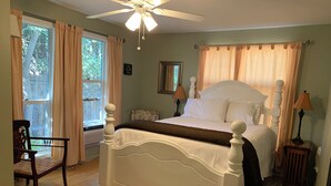 A bedroom with a white four-poster bed, a wooden chair, a small table with a lamp, and a ceiling fan.