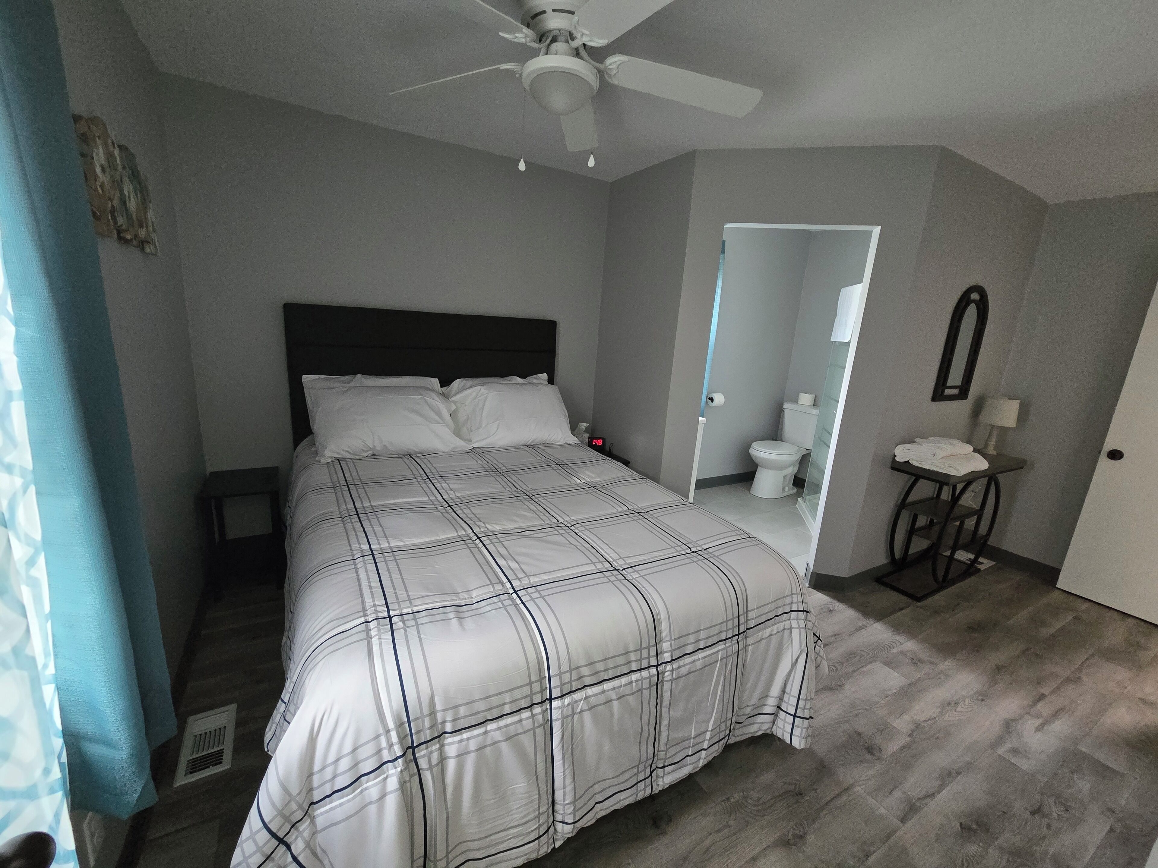 A bedroom with a bed, a ceiling fan, a bathroom with a toilet, and a small table with a lamp.