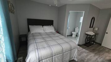 A bedroom with a bed, a ceiling fan, a bathroom with a toilet, and a small table with a lamp.