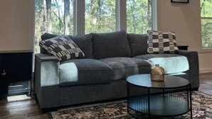 A modern living room with a grey sofa, a patterned throw pillow, a round coffee table, and a large window with a view of trees.