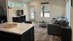 A spacious living area with a dark sofa, a coffee table, and a TV. There is a sign above a door that reads "The Gathering Place."