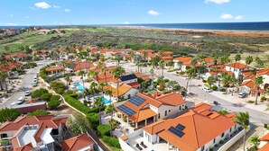 Een kustgebied met woonhuizen, rode dakpannen en zonnepanelen, omgeven door groen en een strand.