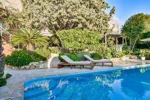 Une piscine bordée de deux chaises longues, entourée de différentes plantes et arbres.