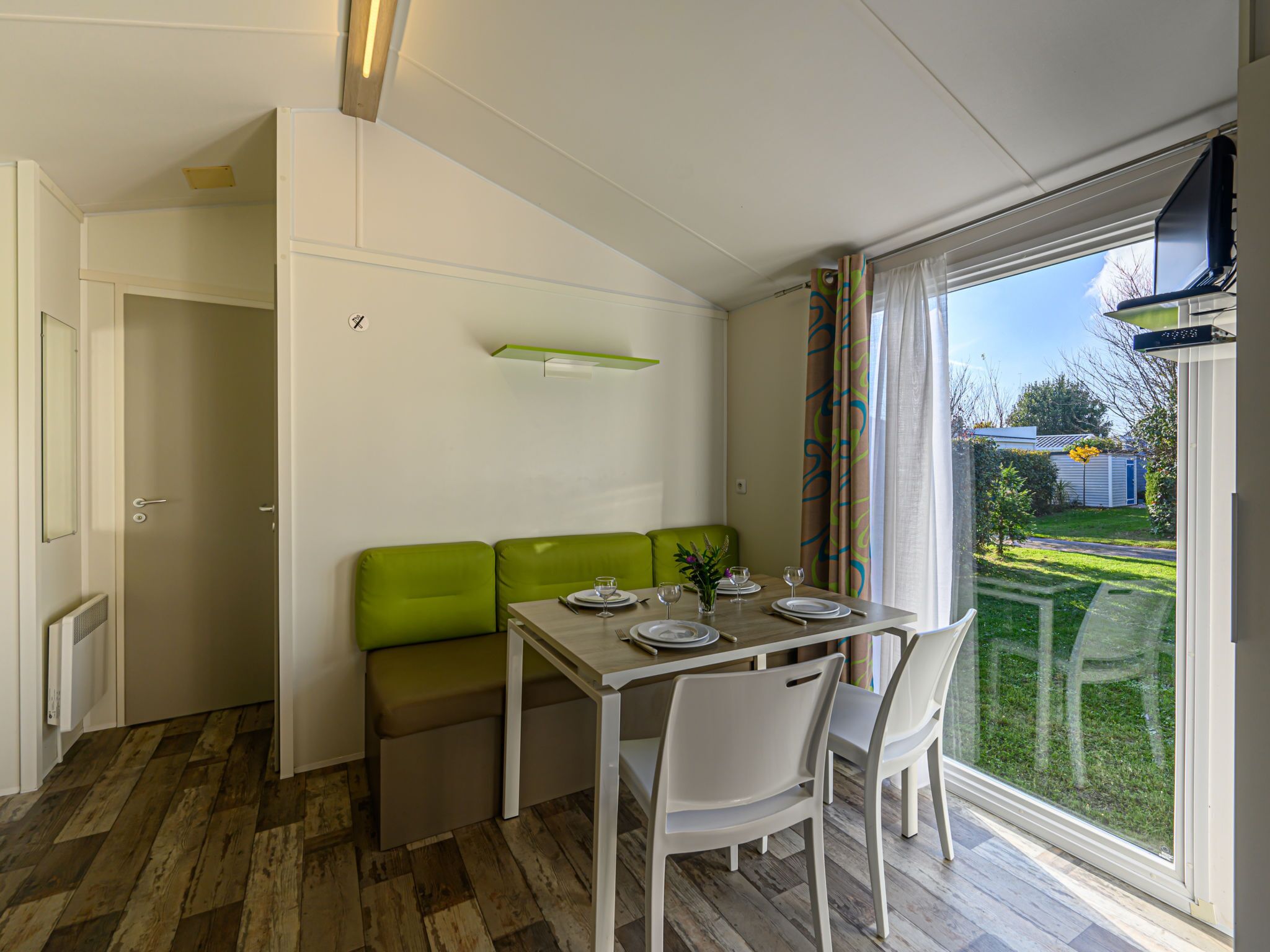 A compact dining area with a table set for four, green cushioned seating, and a view of a garden through a large window.