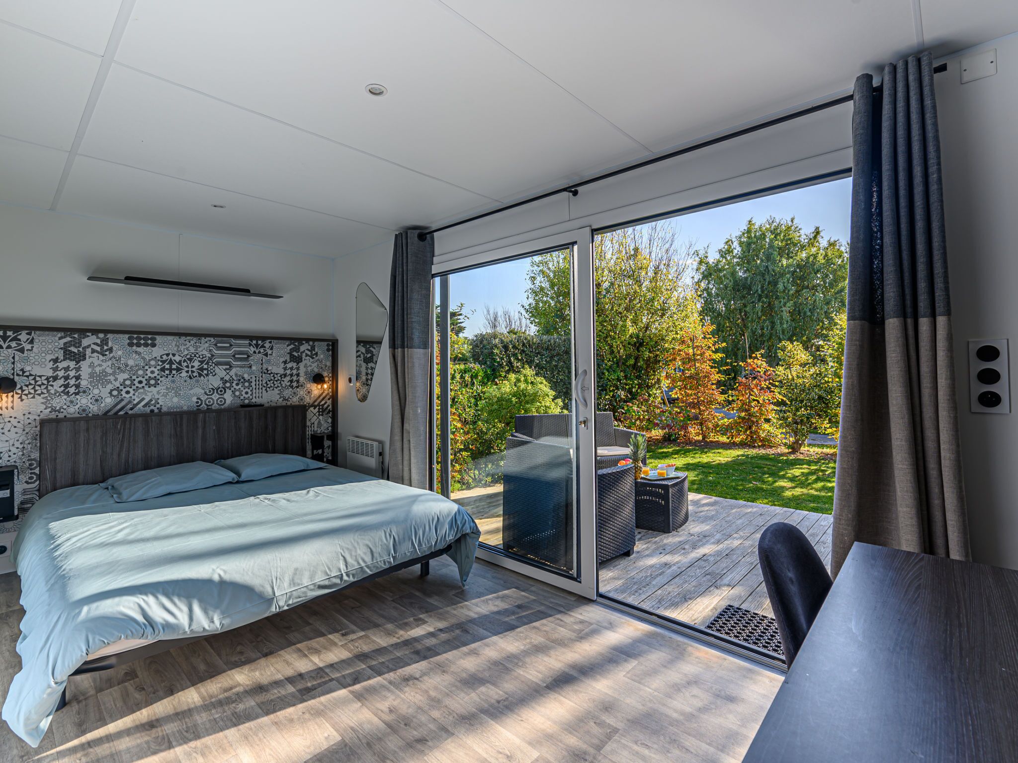 A modern bedroom with a large bed, a sliding glass door to a deck, and a view of a garden.