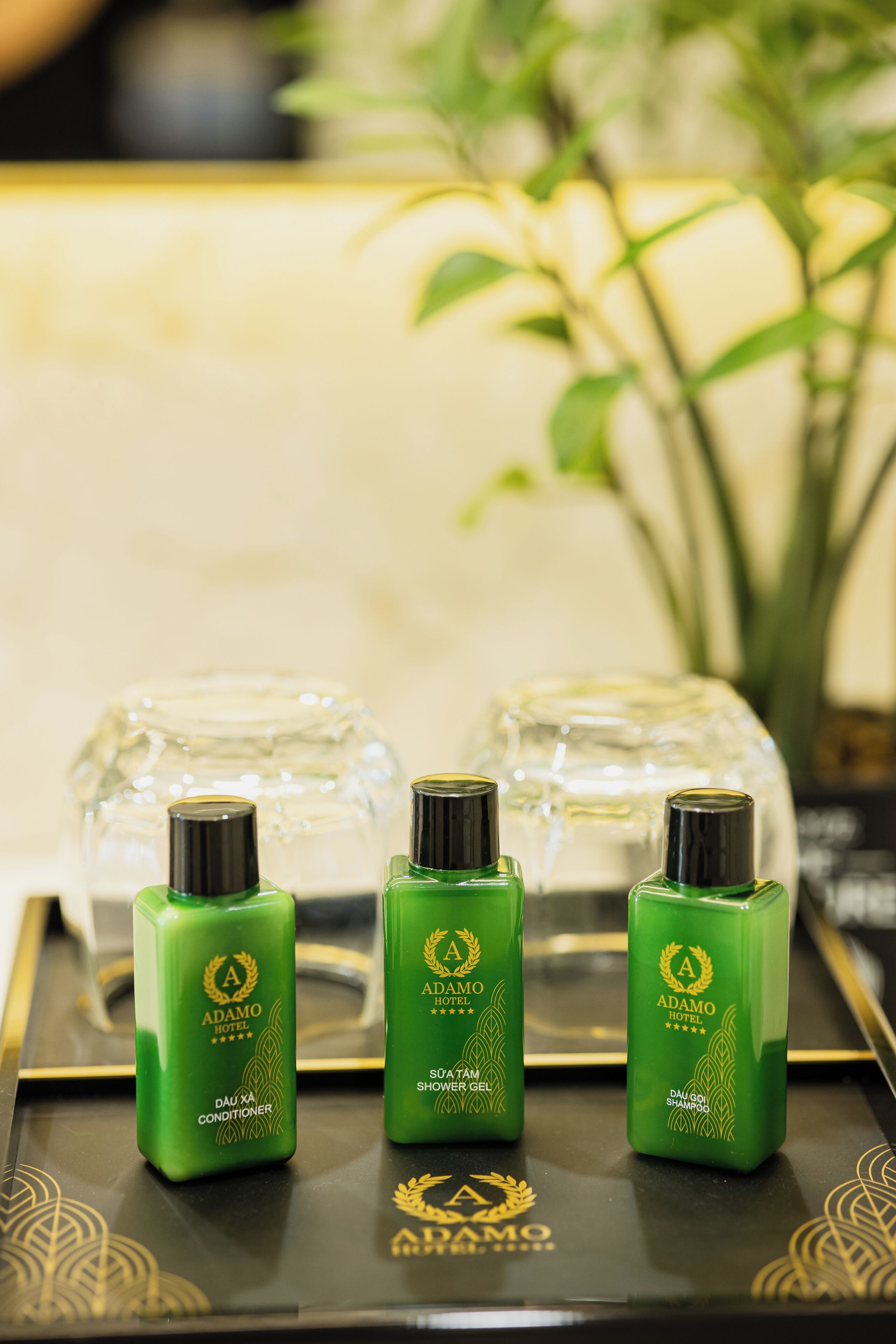Tre grønne flasker Adamo Hotel showergel og balsam på et bakke med gyldent mønster.