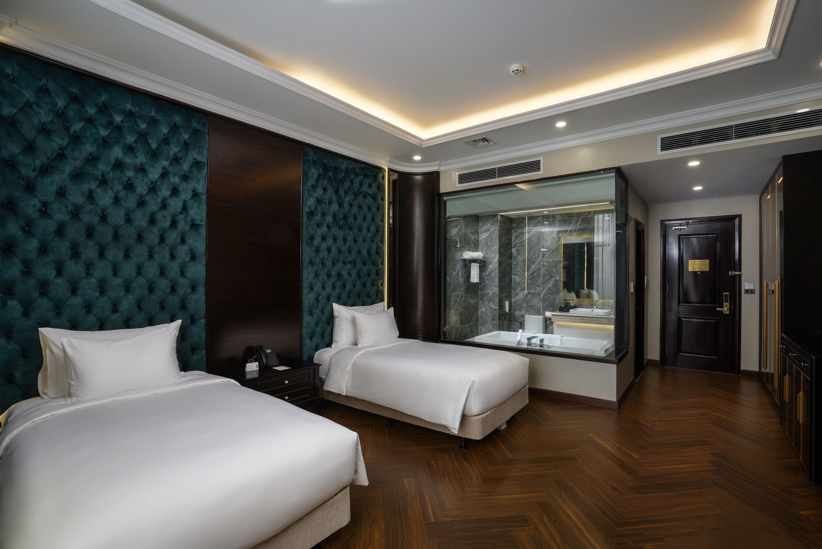 Une chambre d’hôtel avec deux lits, un grand miroir, une salle de bain avec une douche en marbre et un parquet en bois foncé.