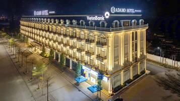 Adamo Hotel features a classic facade with multiple balconies, a well-lit entrance, and a nearby bank.