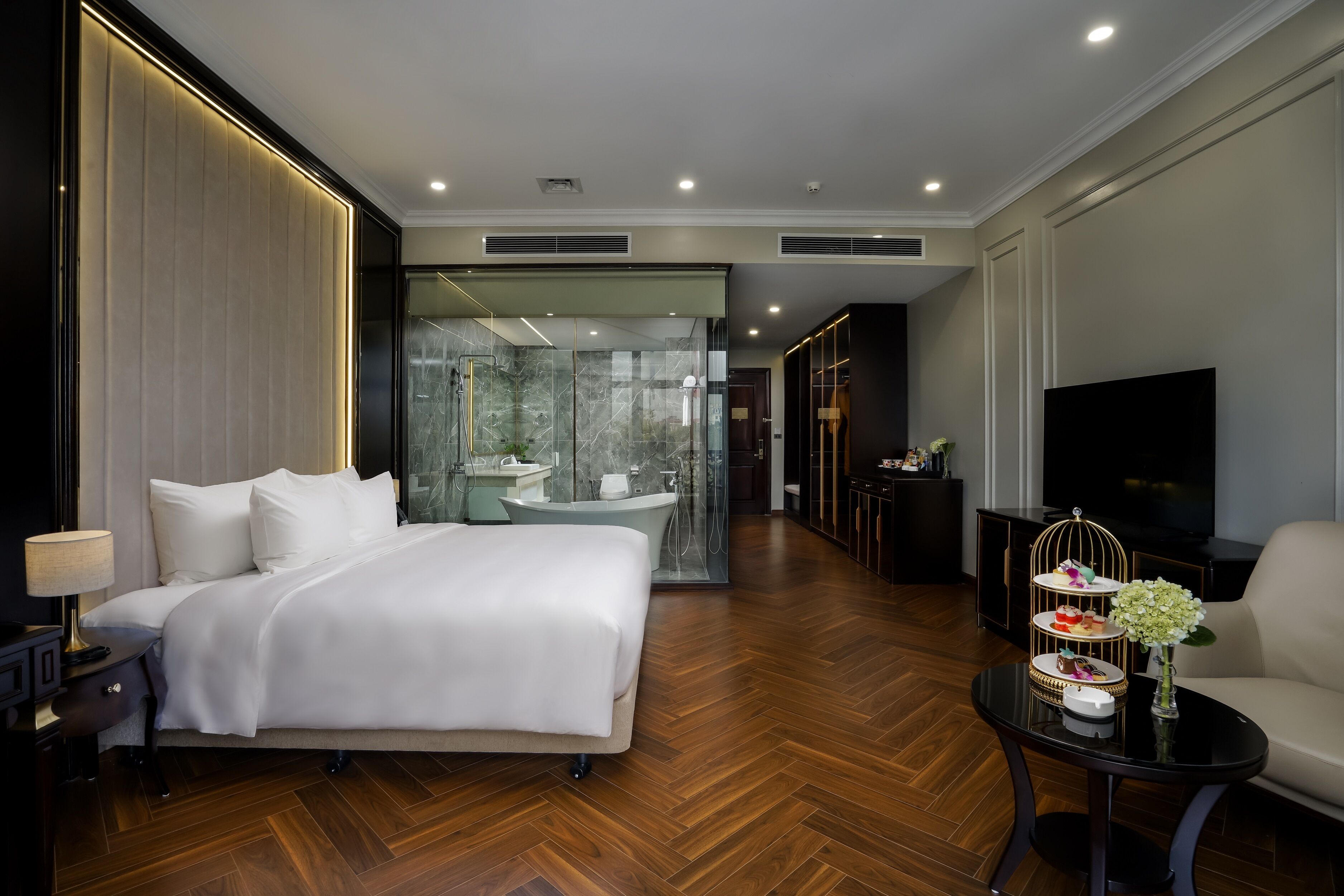 Habitación de hotel moderna con una cama grande, un baño con paredes de vidrio, un televisor de pantalla plana y una pequeña mesa con un objeto decorativo.