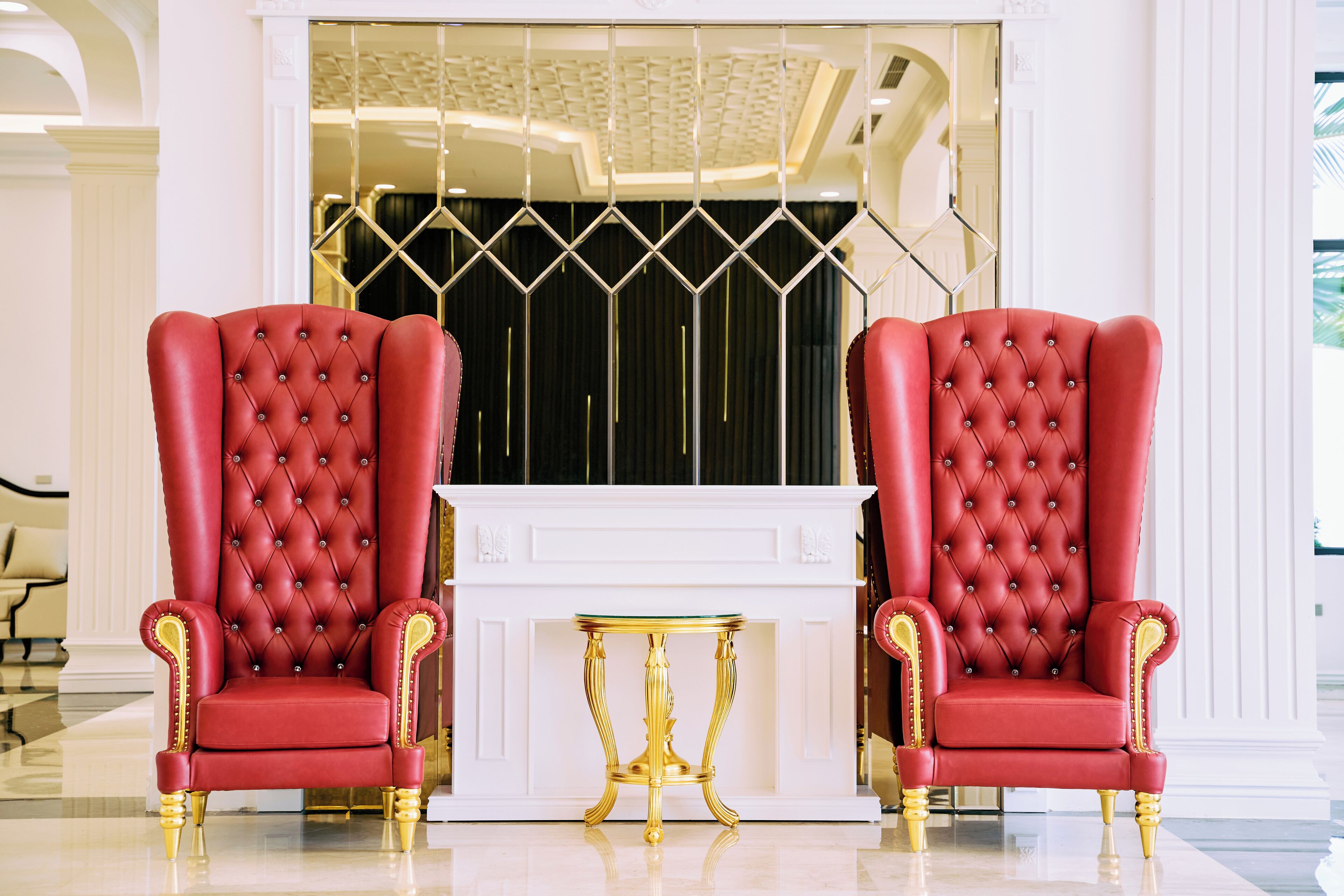 Two red upholstered chairs with gold accents, a gold table, and a white console table in a room with a white fireplace and decorative ceiling.