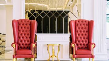 Two red upholstered chairs with gold accents, a gold table, and a white console table in a room with a white fireplace and decorative ceiling.