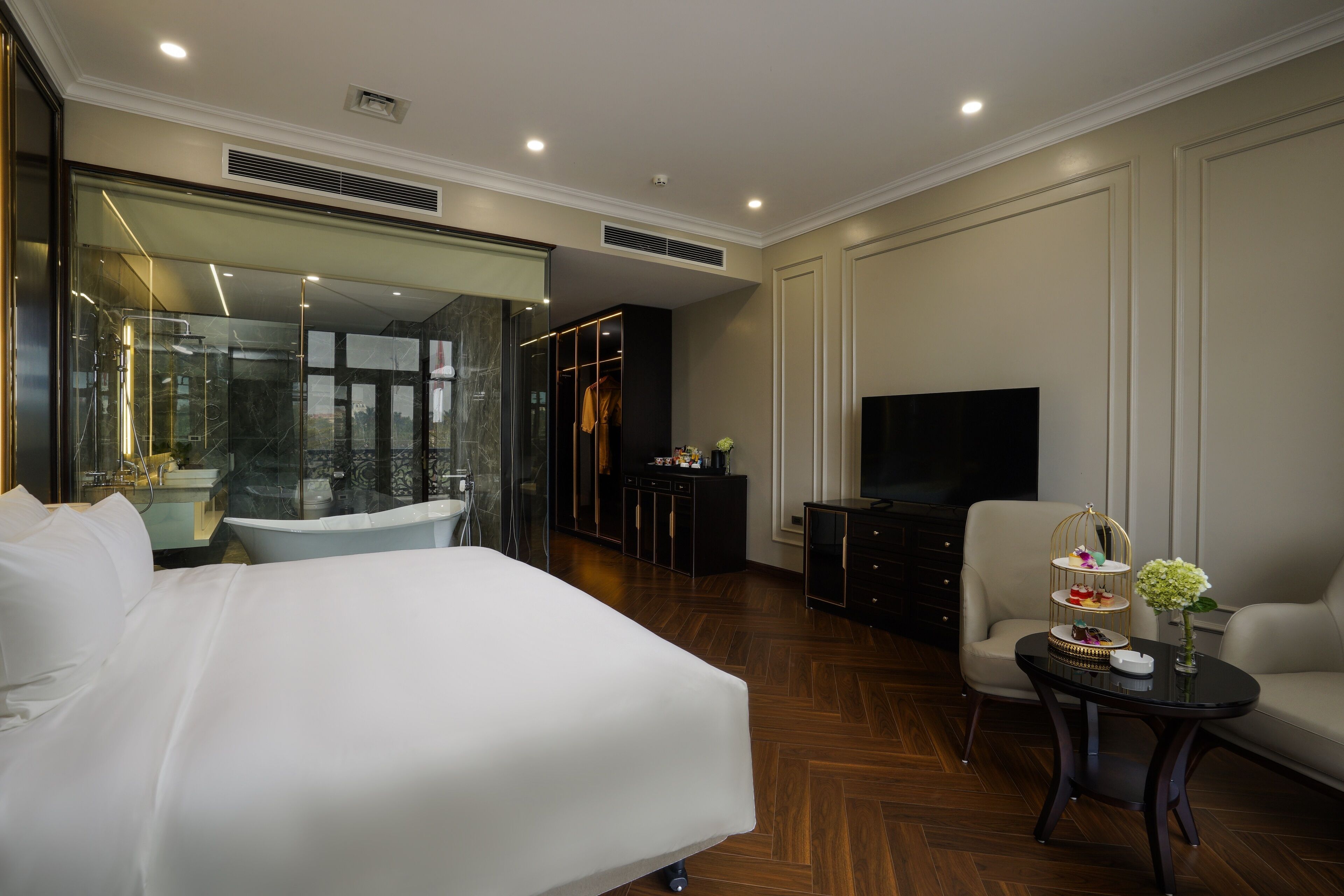 A hotel room with a large bed, a seating area with a sofa and armchair, a small table with a vase, a television, and a glass-enclosed bathroom.