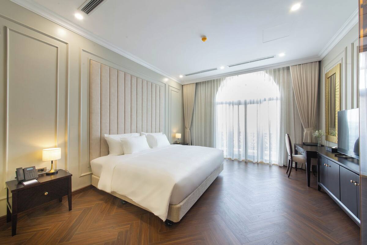 A hotel room with a large bed, a nightstand with a phone and lamp, a desk with a chair, and a large window with curtains.