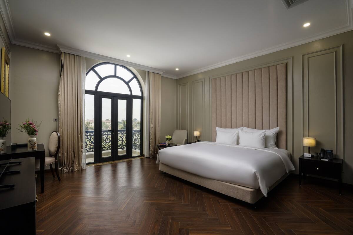 A spacious bedroom with a large bed, a desk with a chair, a nightstand with a lamp, and a view through a large window.