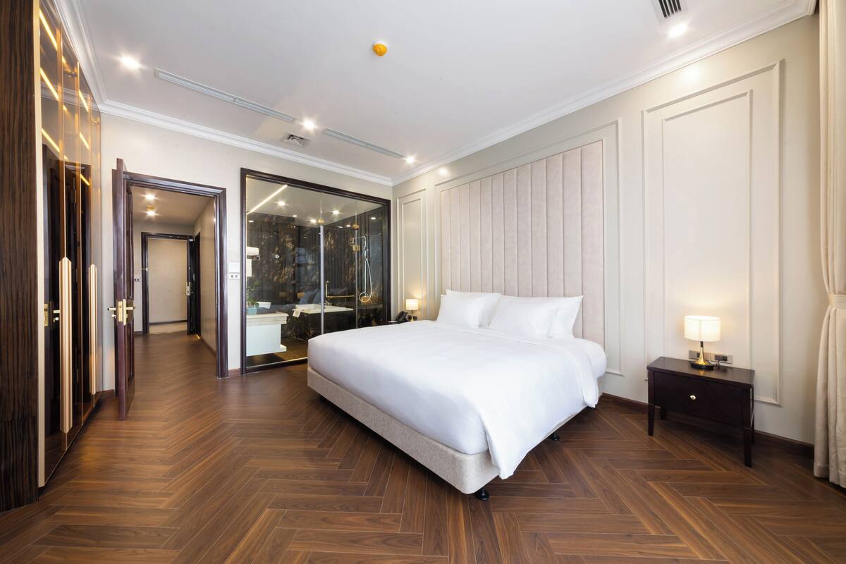 A spacious bedroom with a large bed, a bedside table with a lamp, a mirror, and a wooden floor.