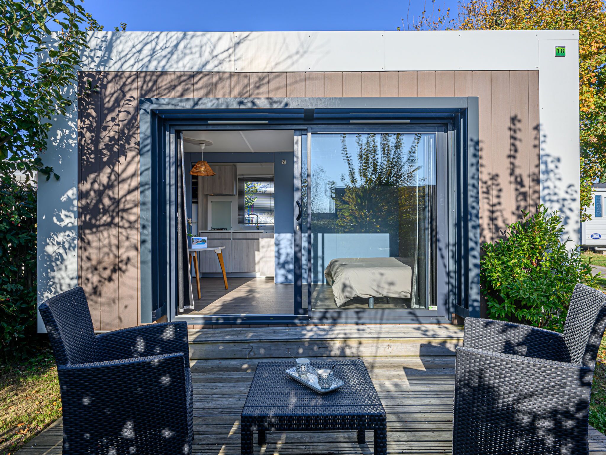 A modern outdoor deck with wicker furniture, a small table, and a view into a bedroom with a bed and a window.