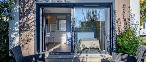 A modern outdoor deck with wicker furniture, a small table, and a view into a bedroom with a bed and a window.