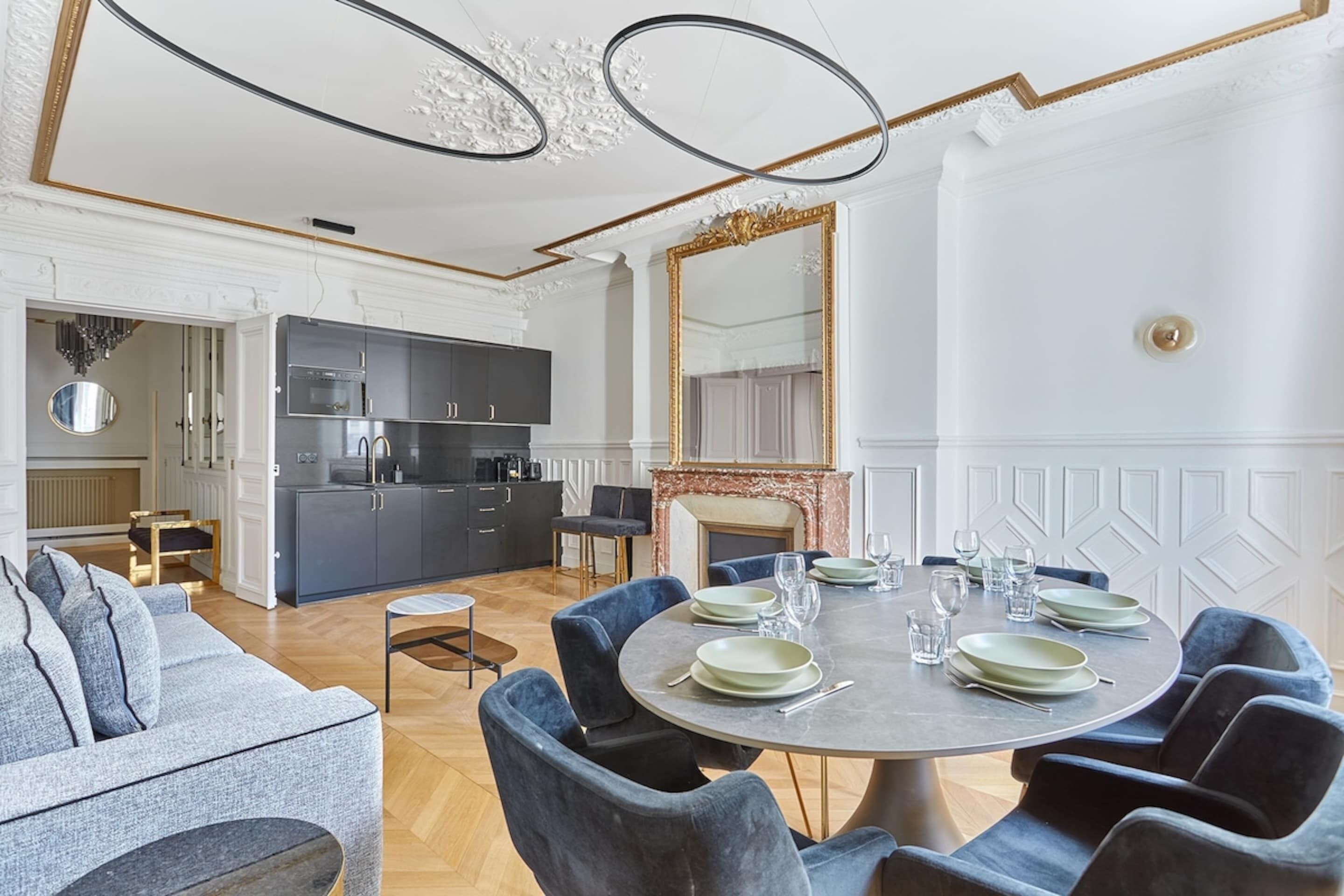 A modern dining room with a round table set for six, a kitchen in the background, and a large mirror above the fireplace.