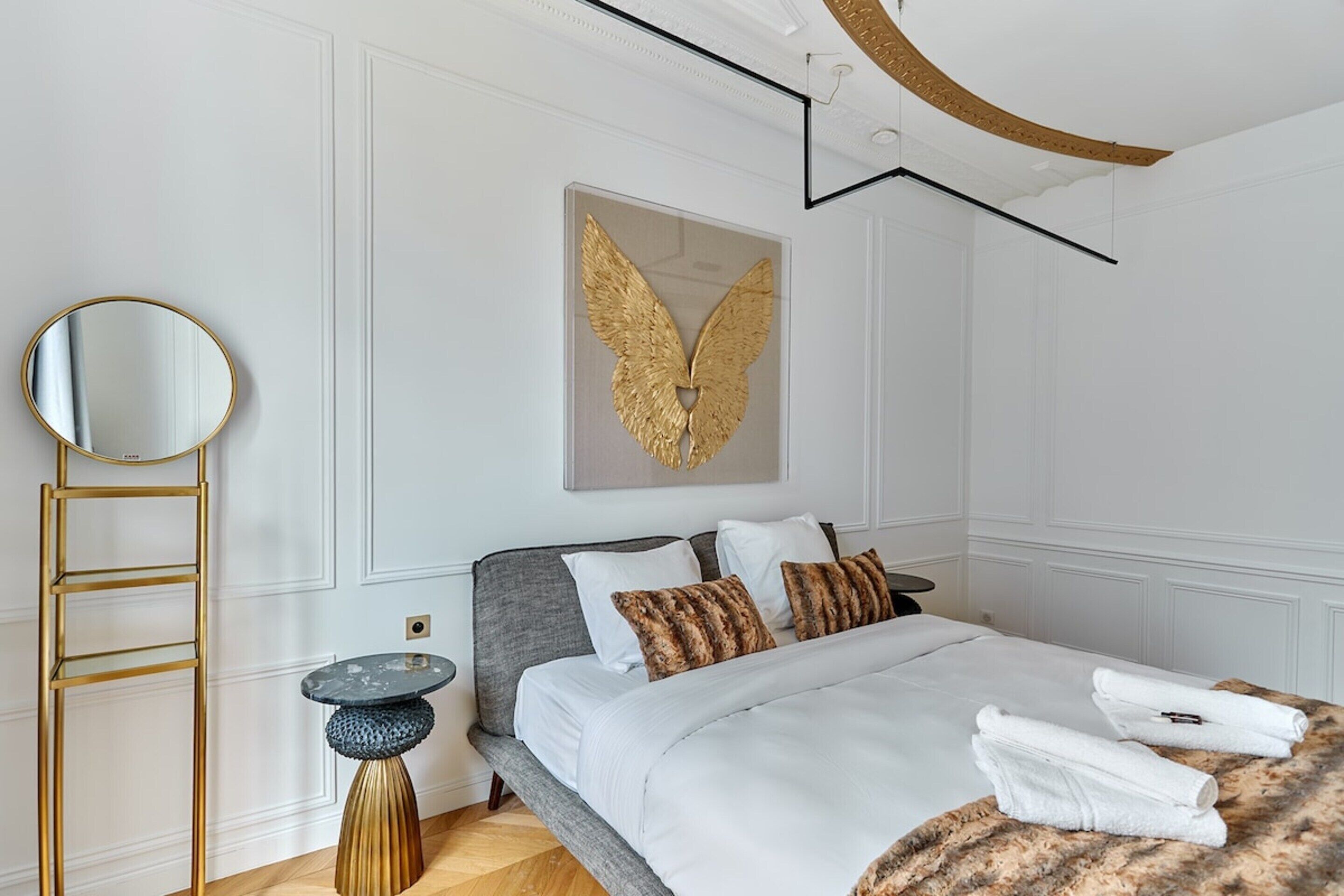 Habitación de hotel con cama, espejo redondo, adorno en la pared y una mesita pequeña y circular.