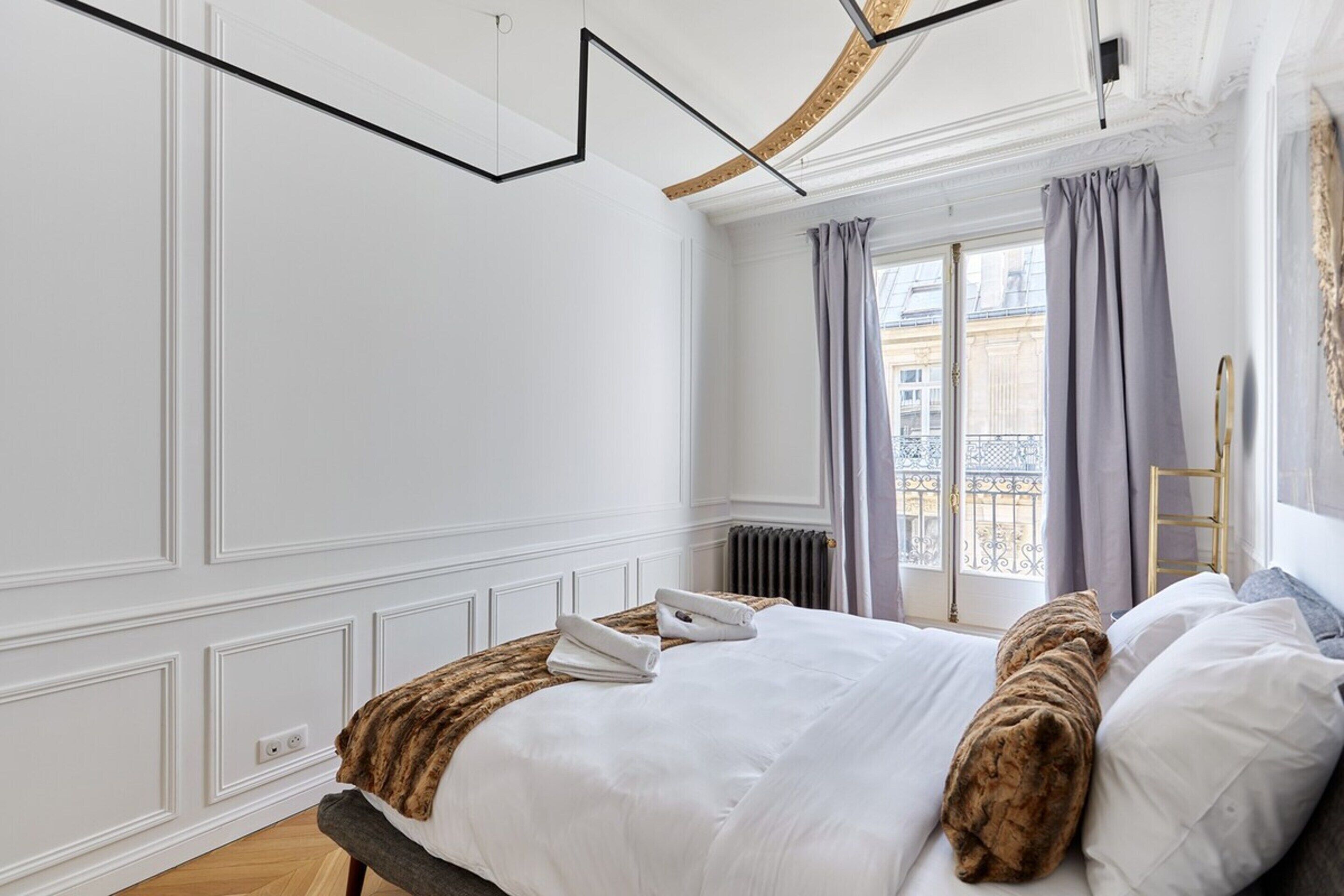 Habitación de hotel con cama, ventana con cortinas, radiador y vistas a edificios.