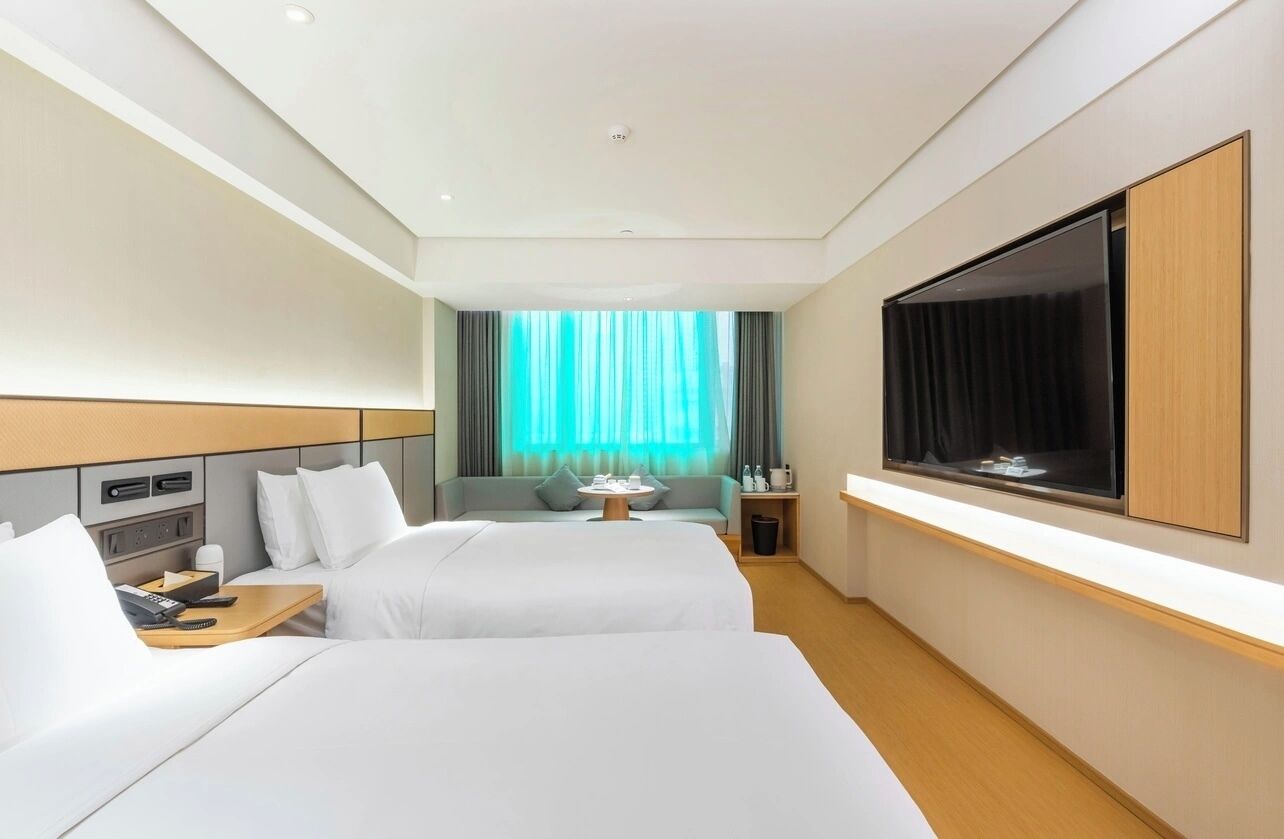 A hotel room with two beds, a TV mounted on the wall, a small table, and a window with curtains.