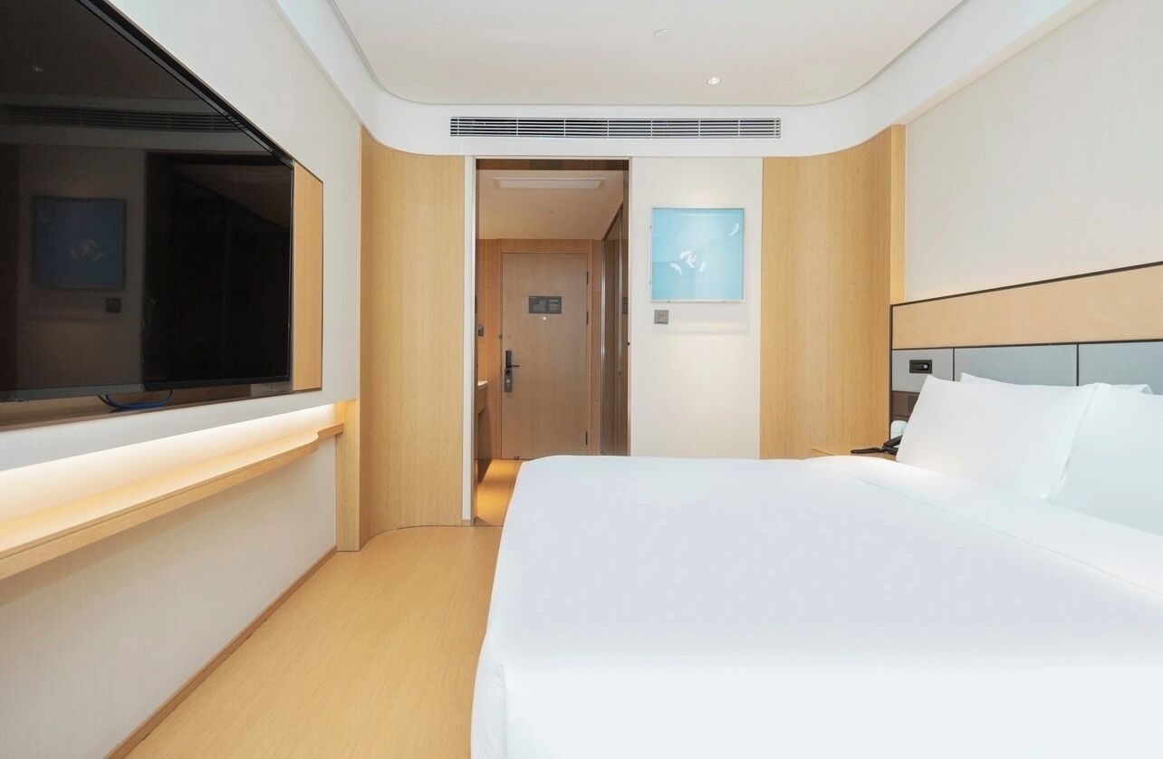 A modern hotel room with a large bed, a bedside table, a window, and a door leading to another room.