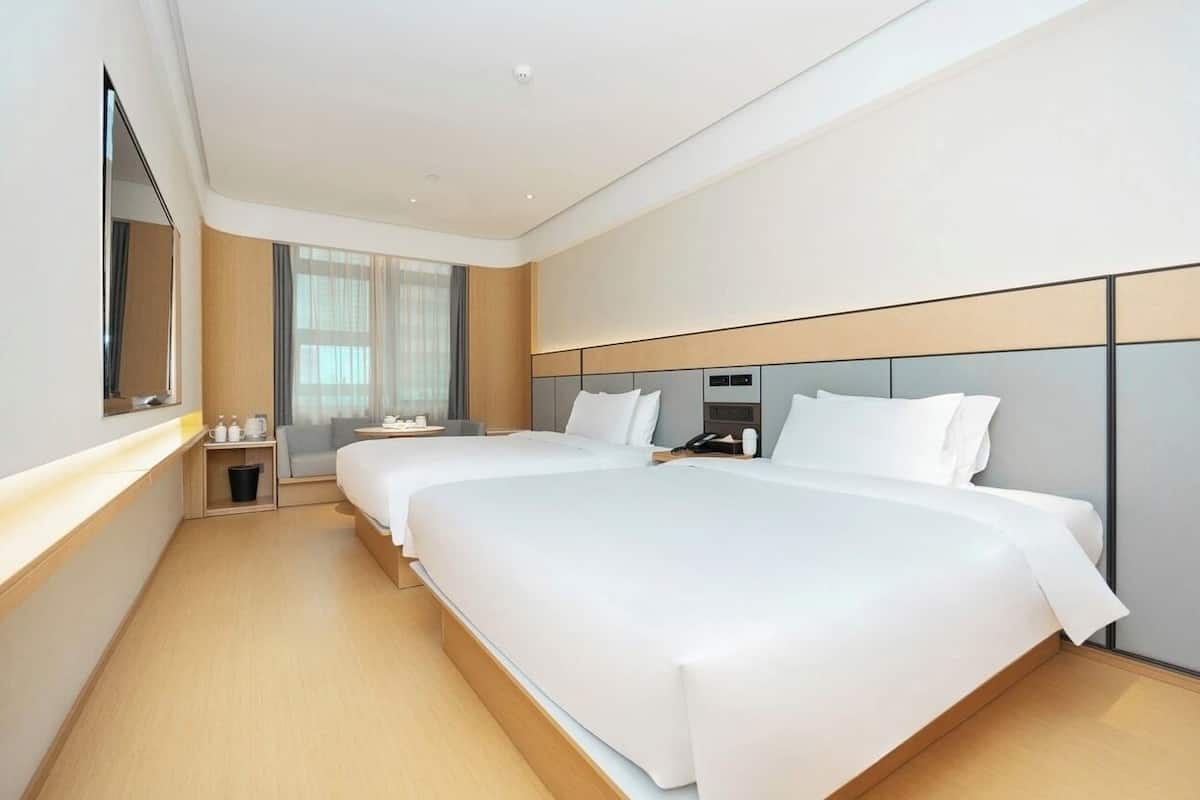 Ein modernes Hotelzimmer mit zwei Betten, einem großen Spiegel, einem Nachttisch und einem Fenster mit Vorhängen.