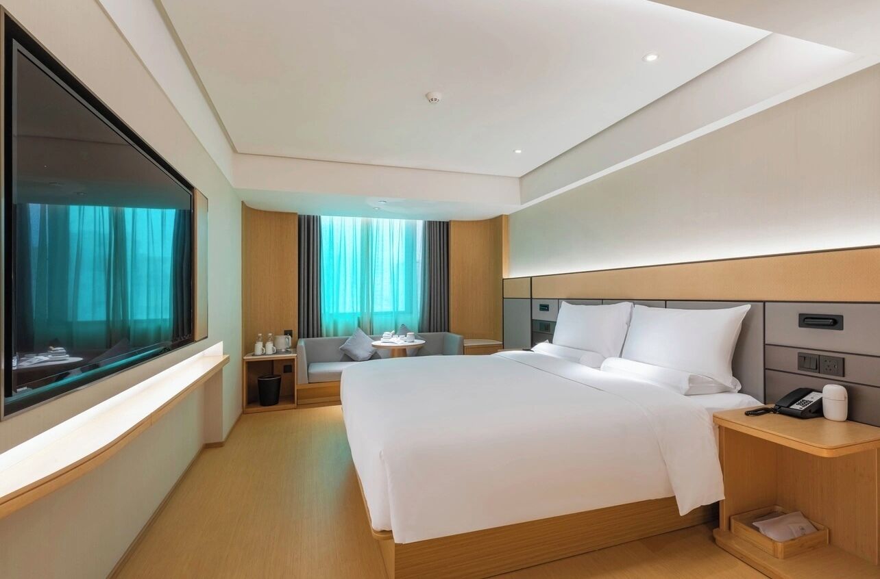 A modern hotel room with a large bed, a bedside table with a phone, a small seating area with a sofa and a table, and a large window with curtains.