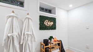 Et badeværelse med to hvide håndklæder hængende på kroge, en træhylde med en varmeblæser, en lille plante og et neon 'Relax'-skilt på væggen.