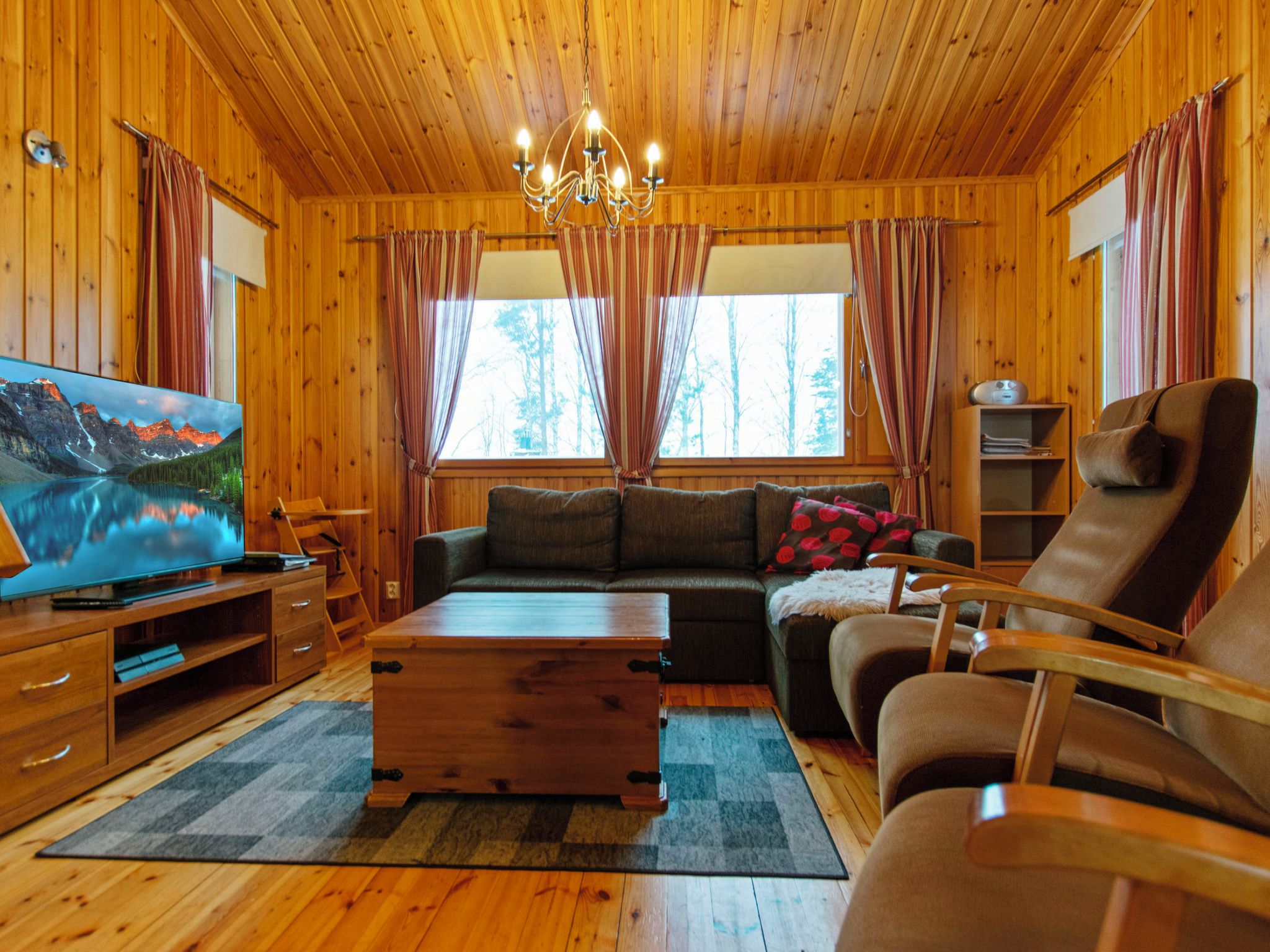 A cozy cabin interior with wooden walls, a large window, a sofa, a chair, a TV, and a wooden coffee table.