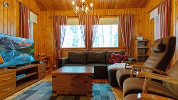 A cozy cabin interior with wooden walls, a large window, a sofa, a chair, a TV, and a wooden coffee table.