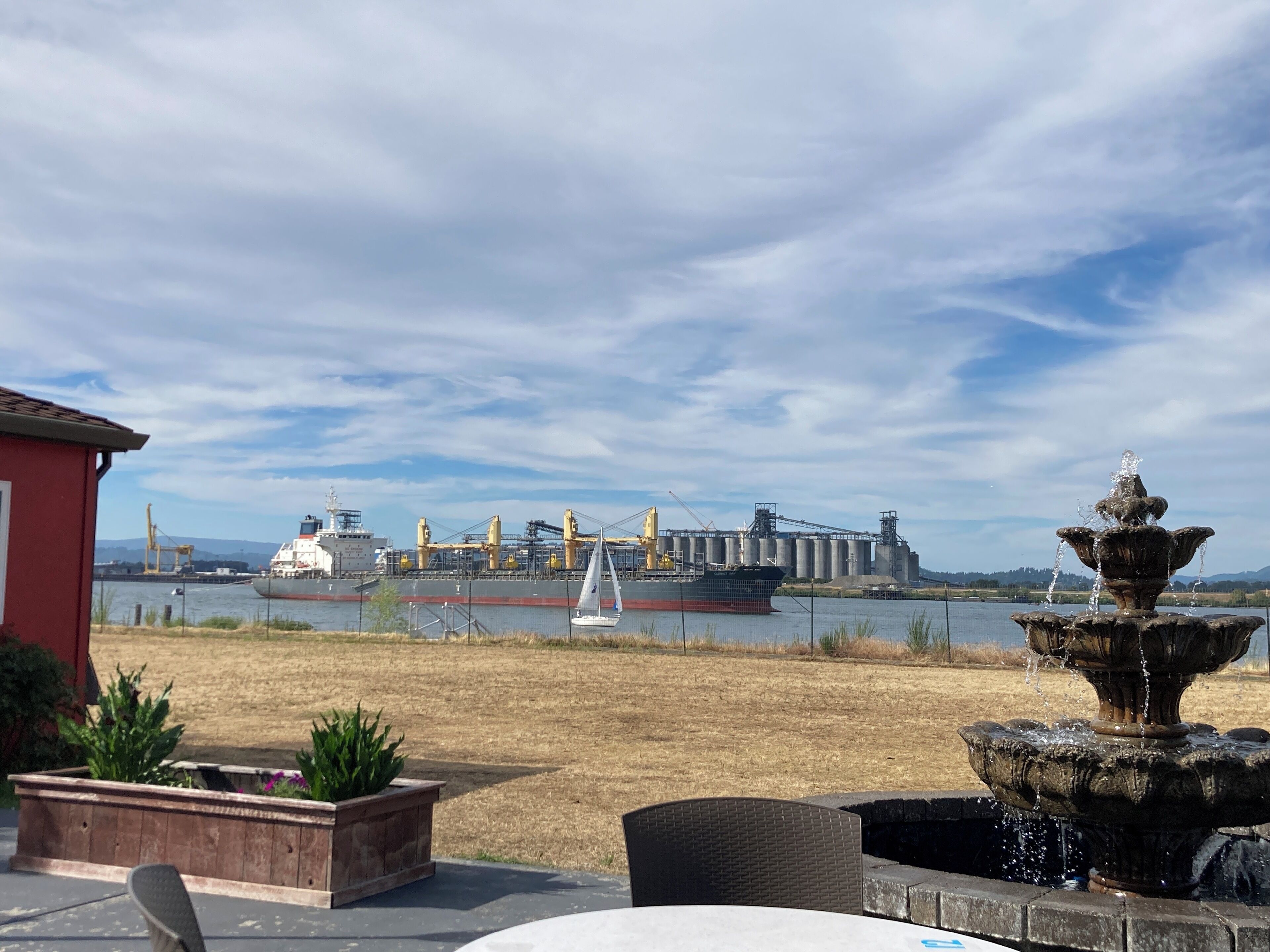A waterfront view with a large industrial facility, a sailboat, and a fountain.