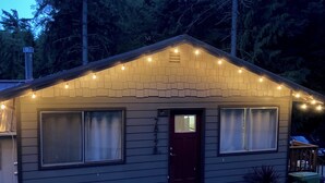 Eine kleine, eingeschossige Hütte mit einer dunklen Vordertür, zwei mit Vorhängen versehenen Fenstern und einer Lichterkette entlang des Daches.