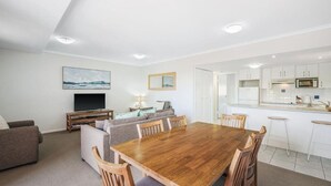 Uma sala de estar moderna com uma mesa de jantar de madeira, sofás, uma televisão e uma cozinha com armários brancos e micro-ondas.