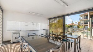 Uma cozinha moderna com azulejos brancos, uma sala de jantar com cadeiras de vime e uma mesa de vidro, e uma varanda com vista para um edifício e áreas verdes.