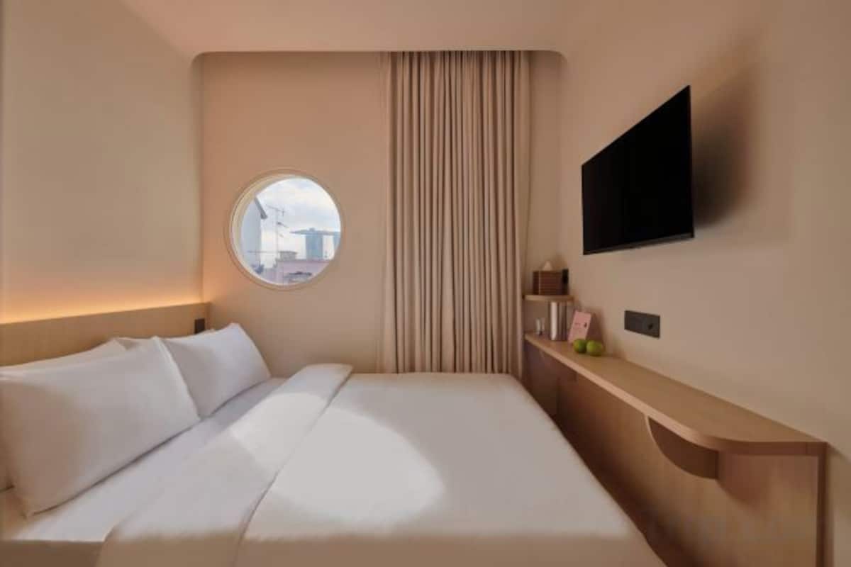 Habitación de hotel con una cama, un ventanal redondo, un televisor y una mesita de madera.