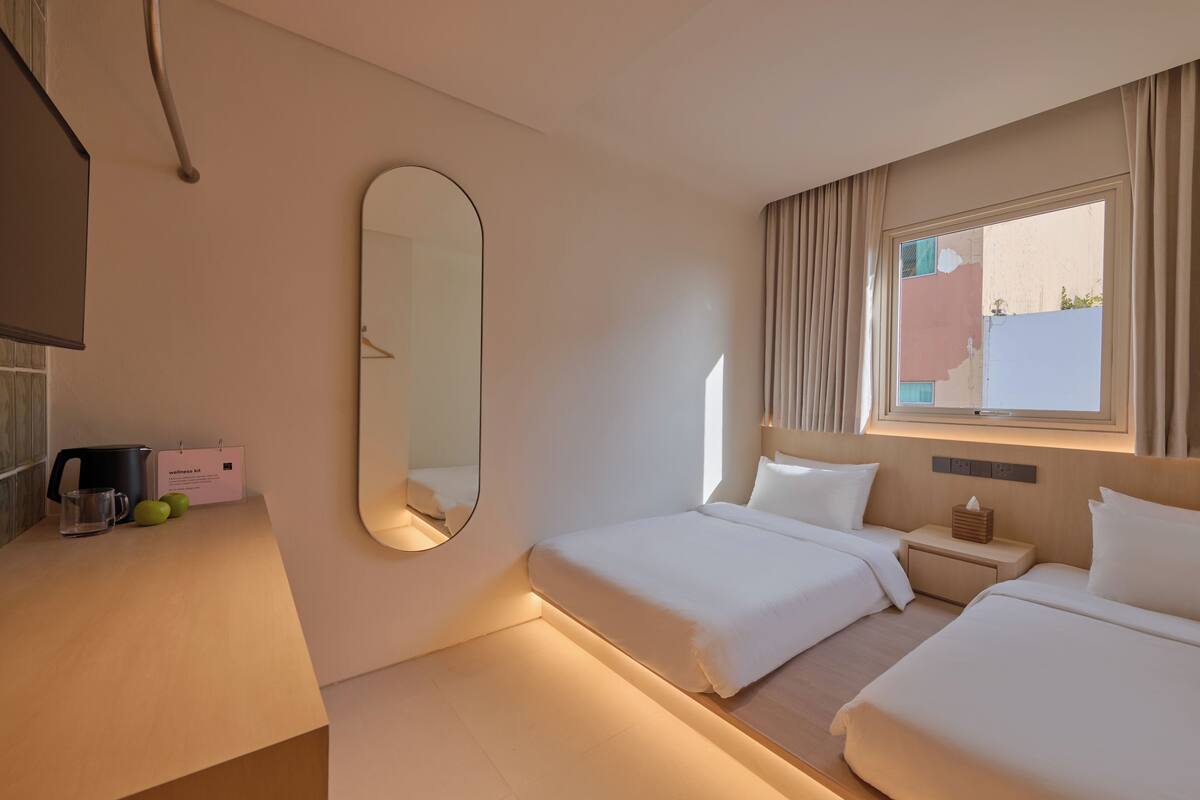 Hotellihuone, jossa on kaksi sänkyä, yöpöytä, peili, ikkuna verhoilla sekä työpöytä, jossa on vedenkeitin ja muki.