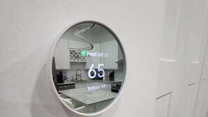 Espejo circular en una pared blanca que refleja una cocina, mostrando una temperatura de 65 grados y una temperatura interior de 66 grados.