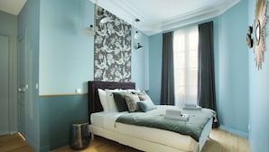 Quarto com cama, parede estampada com flores, janela com cortinas e luminária fixada na parede.