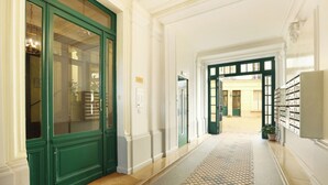 Corredor com portas verdes, piso ladrilhado e vista para um quarto com escrivaninha e cadeira.