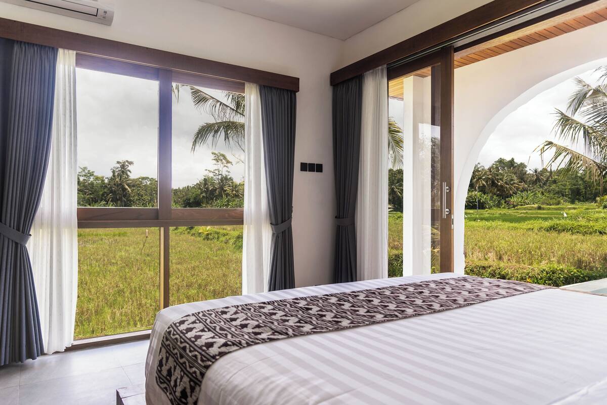 A bedroom with a large bed, a view of a green field, and a sliding glass door.
