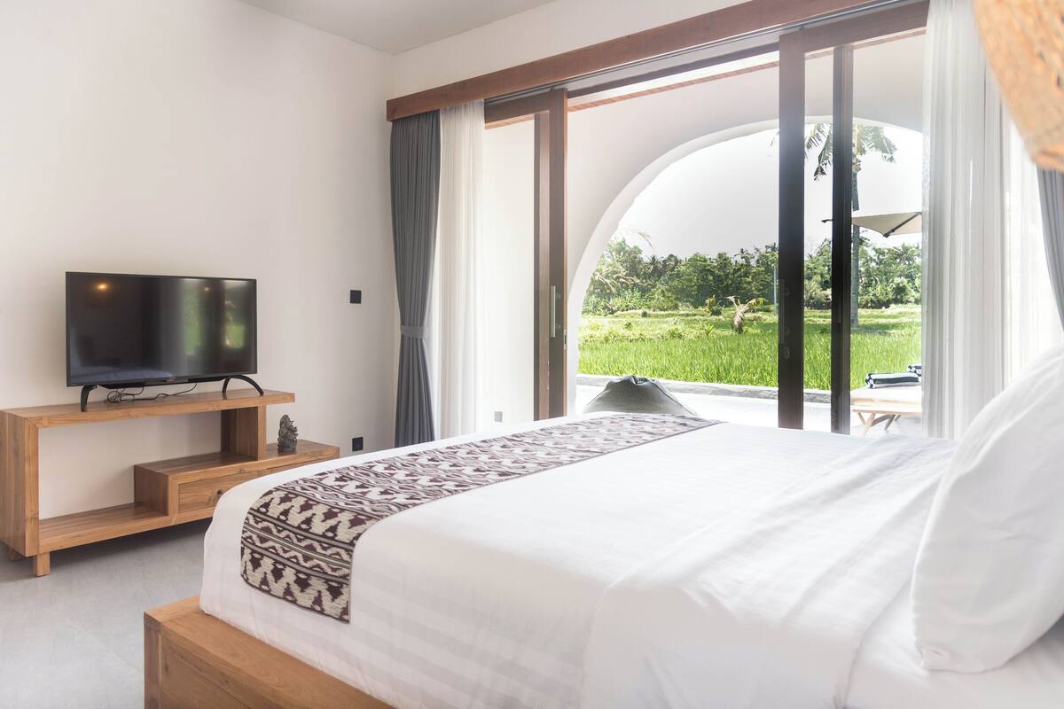 A bedroom with a bed, a TV on a wooden stand, and a view of a green landscape through large windows.