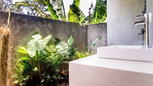 Een badkamer met een wit wastafel, een douche met een handdouche, en een betonnen muur met tropische planten.