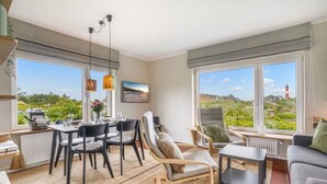 A spacious living room with a dining area, large windows with a view of greenery and a lighthouse, and modern furniture. - Haus Hildebrand - "Relax and feel good in Haus Hildebrand!" (Hörnum)