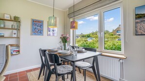 A dining area with a table set for four, chairs, a vase with flowers, and a view of a beach through large windows. - Haus Hildebrand - "Relax and feel good in Haus Hildebrand!" (Hörnum)