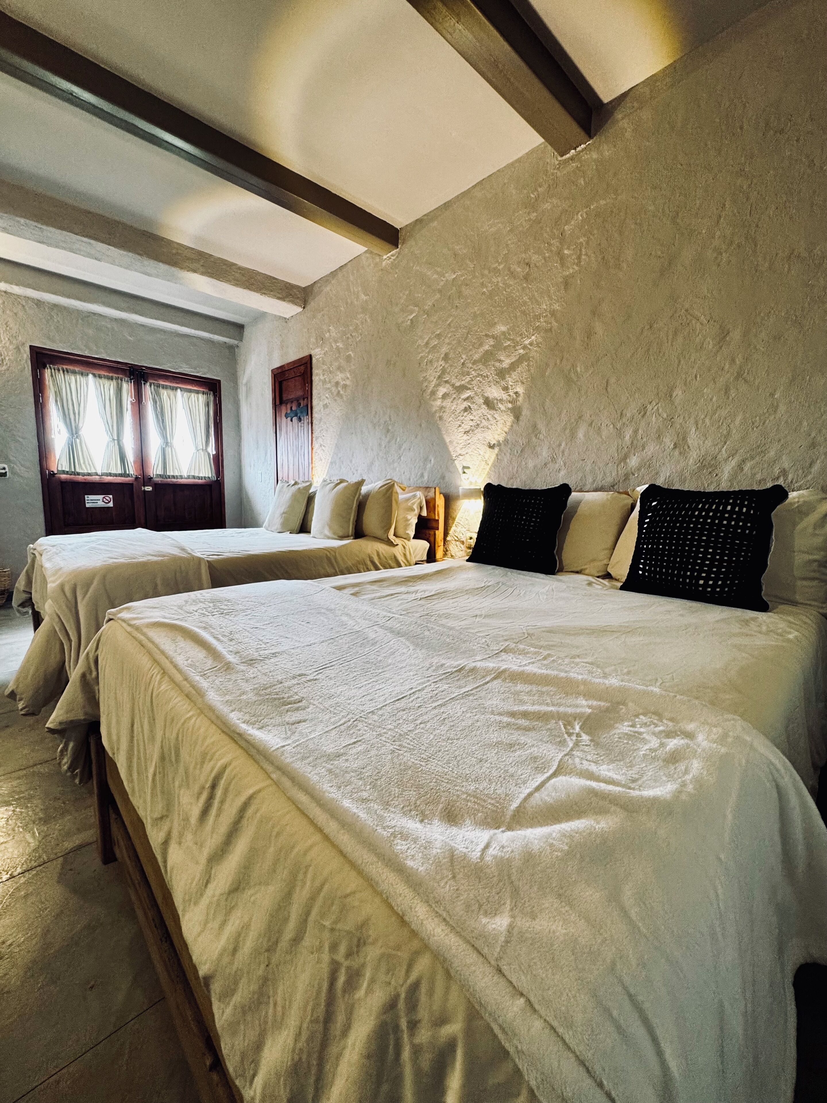 A hotel room with two beds, a wooden ceiling, and a large window with curtains.