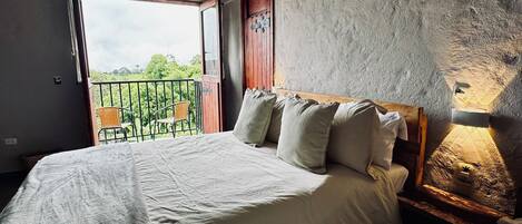 A bedroom with a large bed, a bedside table, a wooden headboard, a window with a view of greenery, and a door.